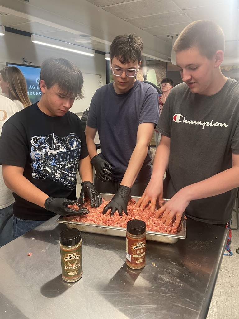 Three students mix seasoned ground pork by hand in a large metal pan, with sausage seasoning containers on the table in front of them.
