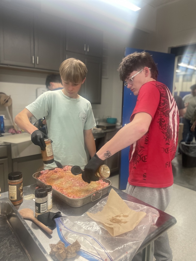 Two students sprinkle seasoning over a large pan of ground pork while preparing sausage in class.