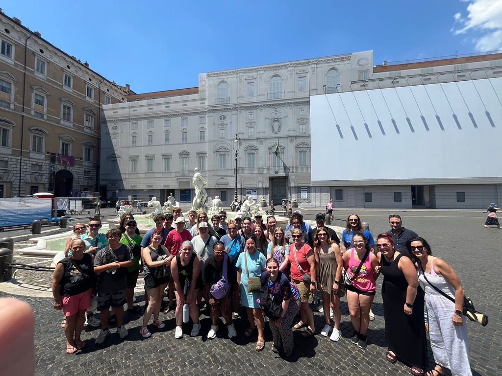 A group of Savannah High School students and chaperones pose in a sunny plaza in Rome near a fountain and historic buildings.