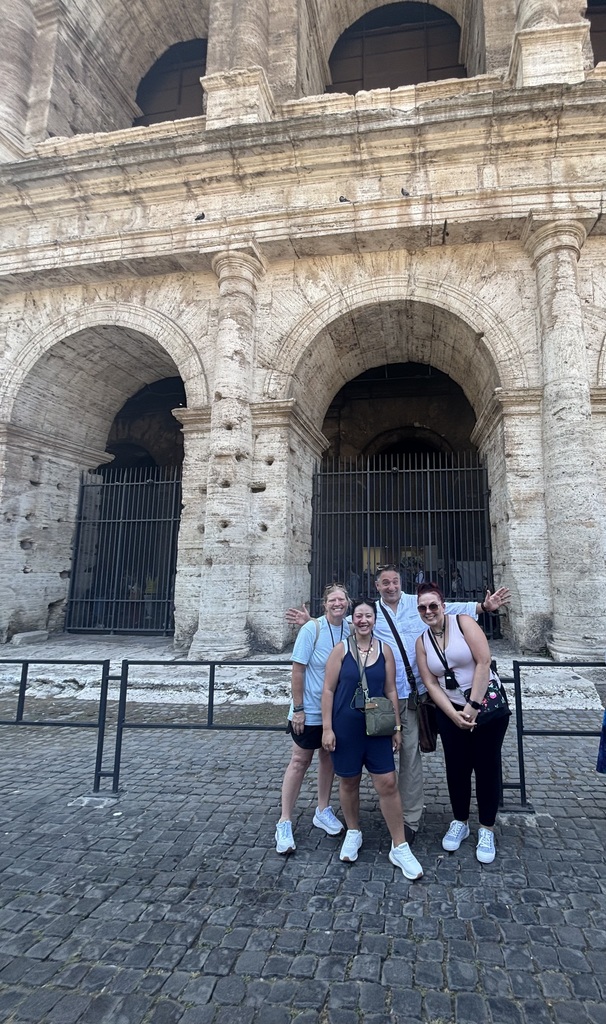 Four travelers smile in front of the Colosseum in Rome, standing on a cobblestone street near the stone arches.