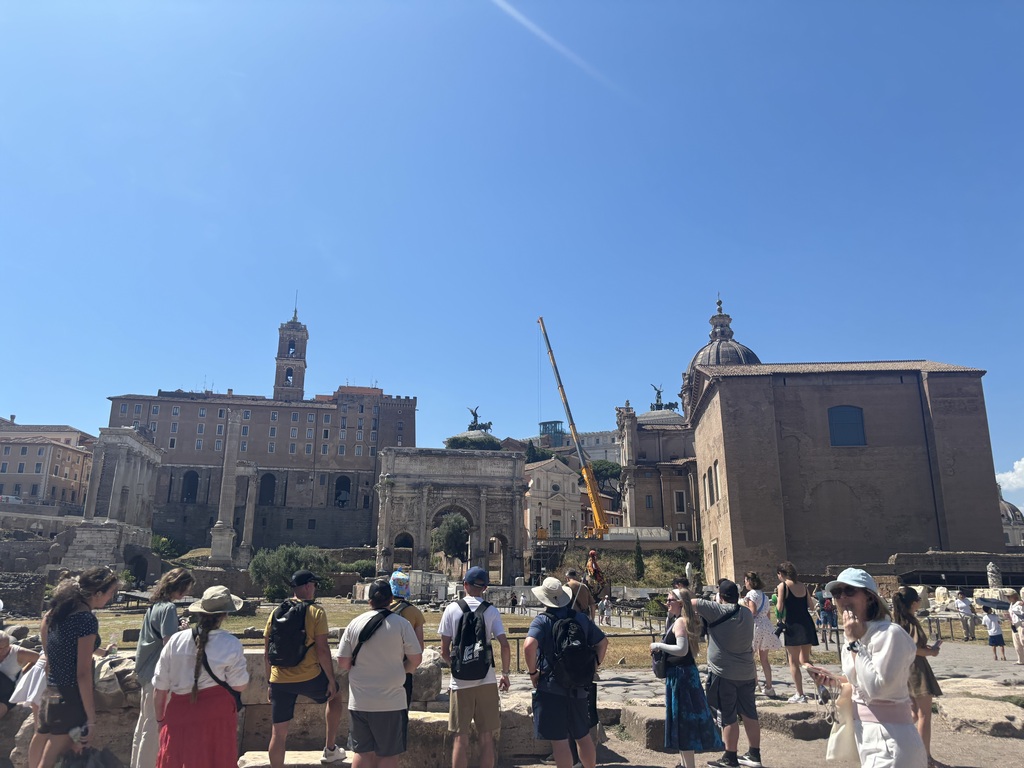 Students explore ancient ruins with historic buildings and arches in the background under a clear blue sky.