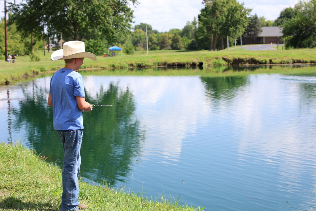 A student wearing a cowboy hat fishes from the grassy bank of a pond on a sunny summer day with trees reflected in the calm water.