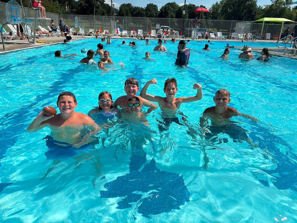 Five smiling children pose together in a community swimming pool while families enjoy a sunny summer afternoon in the background.