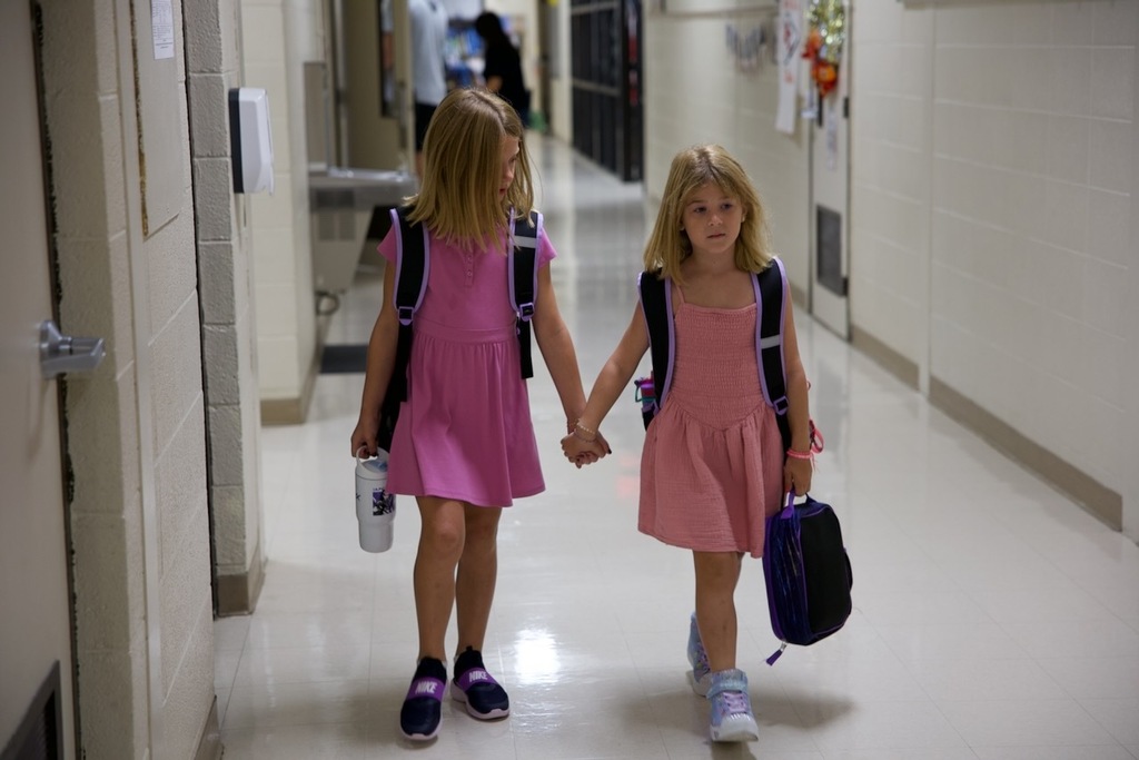 Two young students hold hands as they walk down a school hallway with backpacks and lunchboxes.