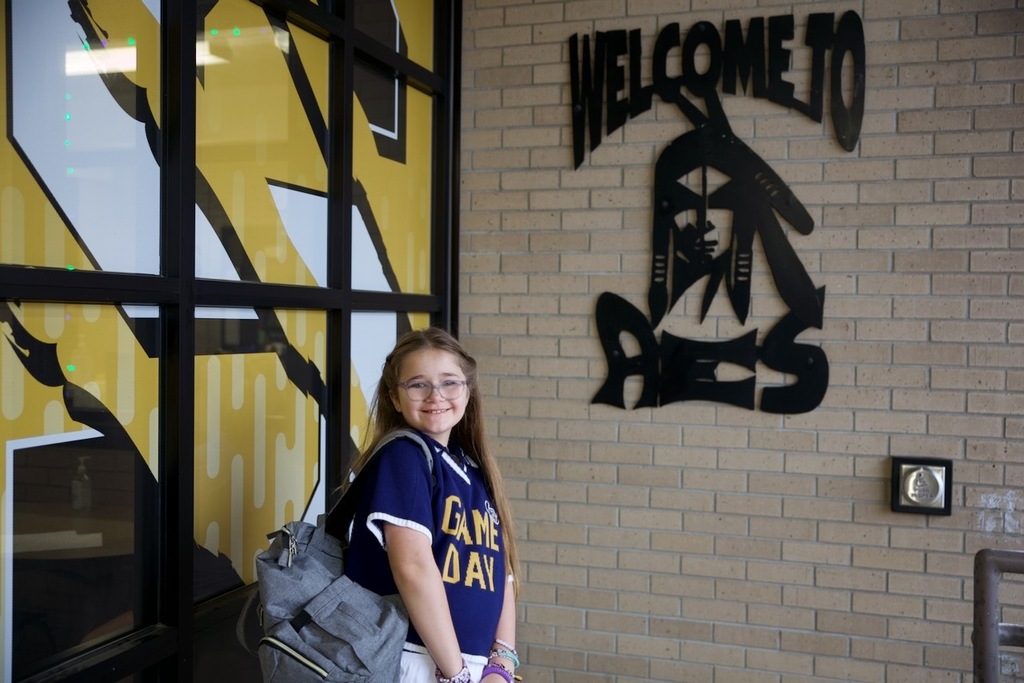 Student smiles with her backpack near a “Welcome To” Savage display on the first day of school.