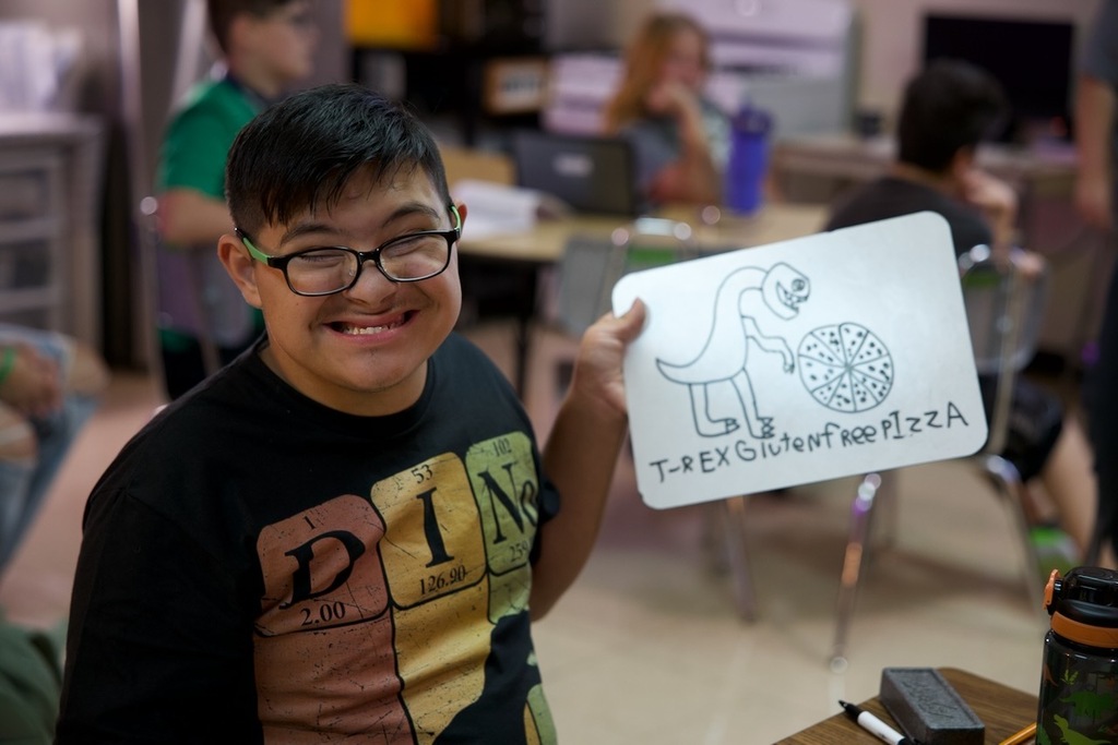 Student proudly holds up his “T-Rex Gluten Free Pizza” drawing while smiling in class.