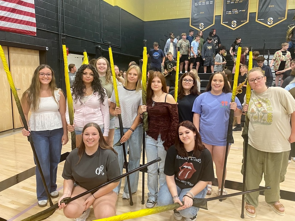 High school color guard members pose with flags in the gym as band students gather behind them.