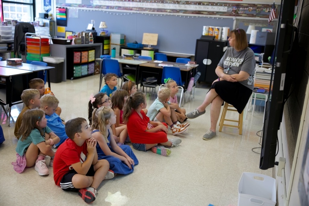 13 first grade students sit on the floor of their classroom and listen to their teacher for circle time