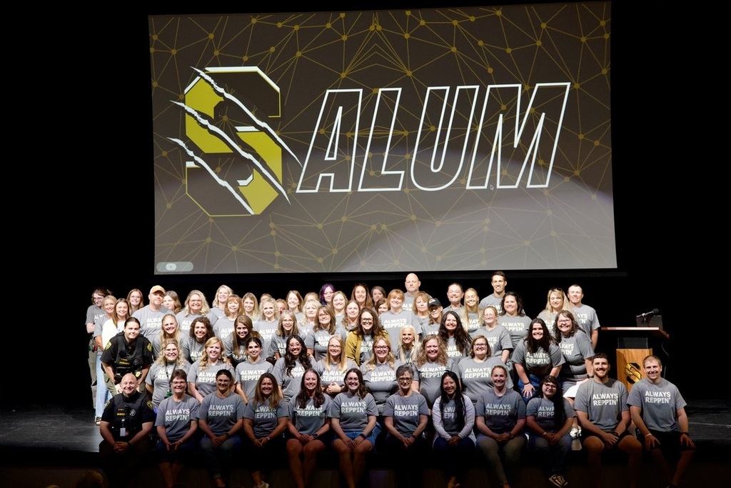 Large group of Savannah R3 staff members who are also district alumni pose together on the Performing Arts Center stage beneath a large “S ALUM” display.