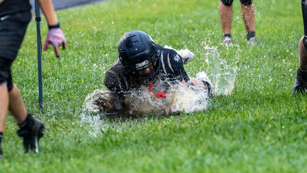 Middle school football player slides through a large puddle on the grass, sending water splashing around him.