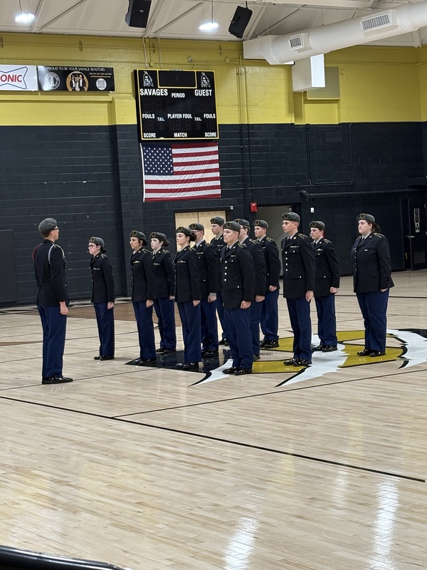 JROTC cadets in uniform stand in formation on Savannah High School gym floor beneath American flag.