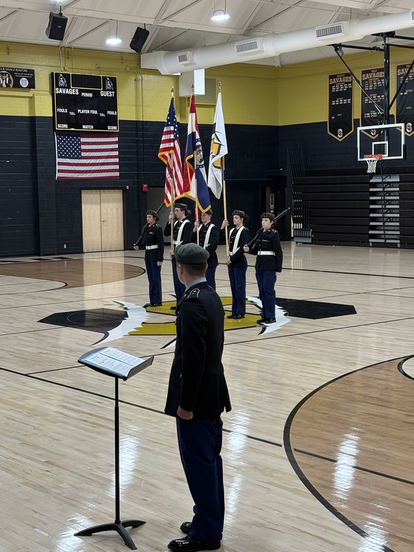 JROTC color guard presents flags in school gym while a cadet reads from a podium in the foreground.