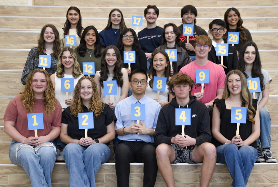 twenty high school students sit on interior stairs holding numbered placards, 1–20