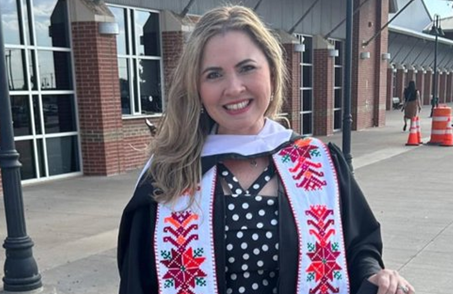 woman in graduation regalia stands outside a brick & glass building, smiling