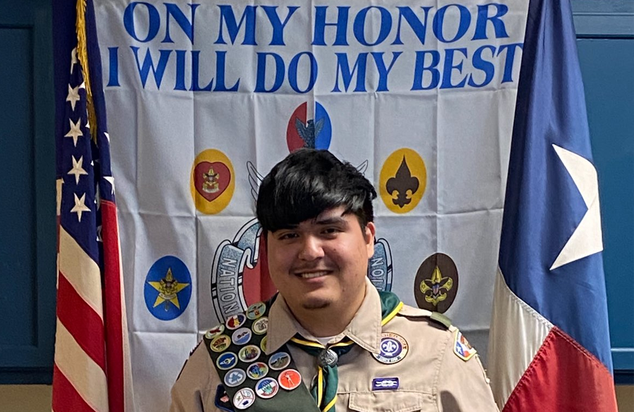 a boy in a boy scouts uniform stands centered between us & texas flags and a banner that reads "on my honor i will do my best"