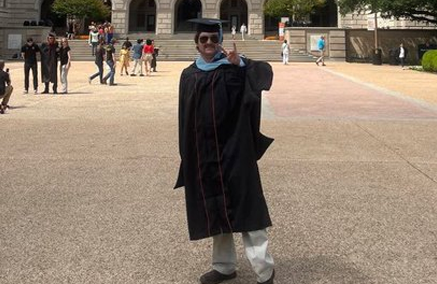 a man in sunglasses & graduation regalia stands before the tower at ut austin extending index & forefingers to signify "hook 'em horns"