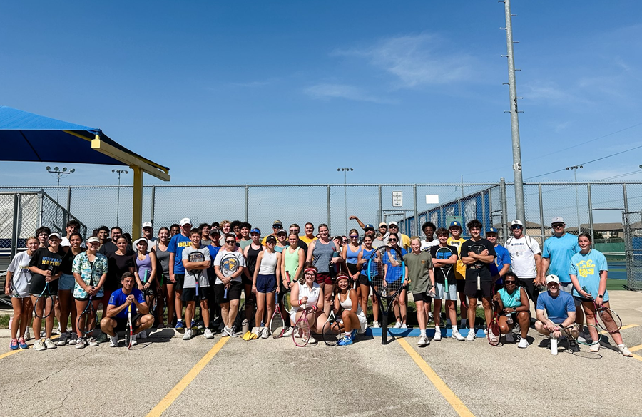 students & teachers with tennis rackets stand before tennis courts beneath a blue sky