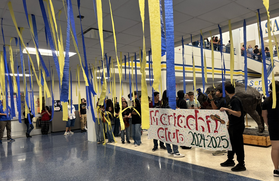students line school hallways holding a banner which reads "mariachi club 2024-2025." blue & yellow streamers hang from the ceiling