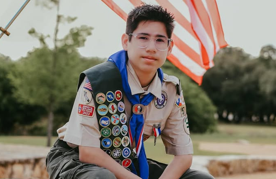 boy in glasses with dark hair in boy scouts uniform sits on a rock with an american flag billowing in the background
