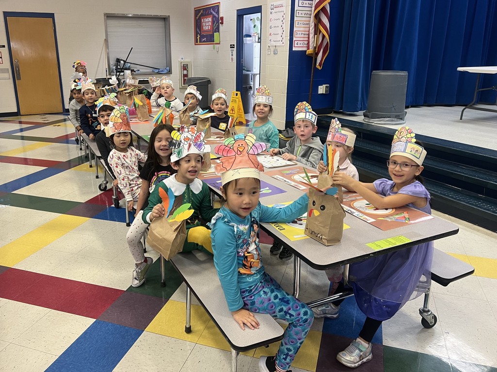 students wearing handmade turkey hats sitting in the cafeteria with handmade thanksgiving placemats in front of them.
