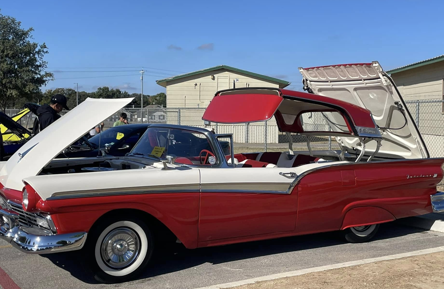 1950s ford fairlane convertible in 2-tone white and red with whitewall tires sits in a high school parking lot with hood & trunk open and roof partially retracted.
