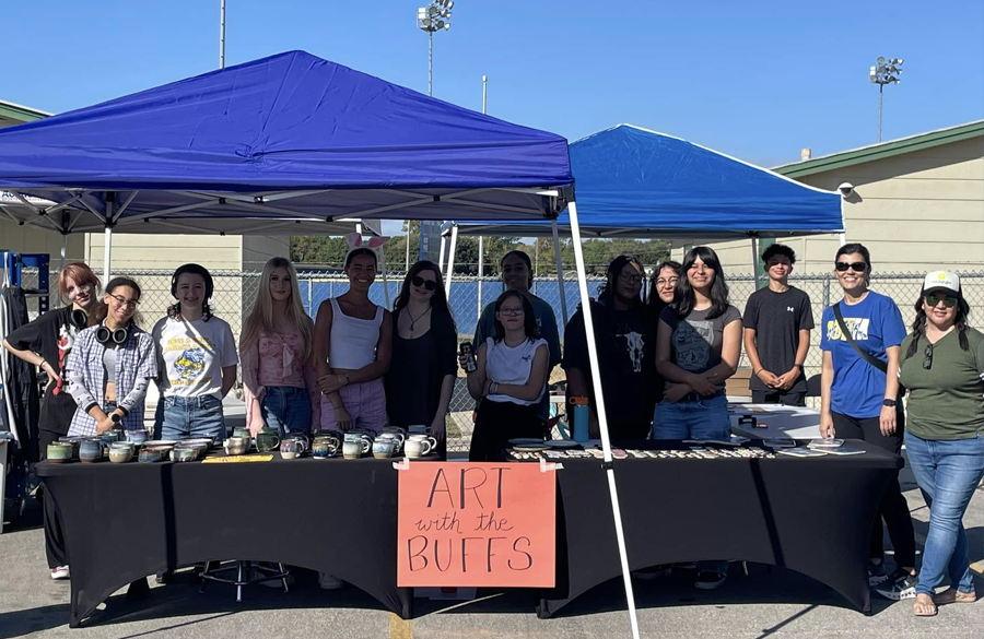 high school students stand behind a long black table and blue canopies. a sign on the table reads "art with the buffs"