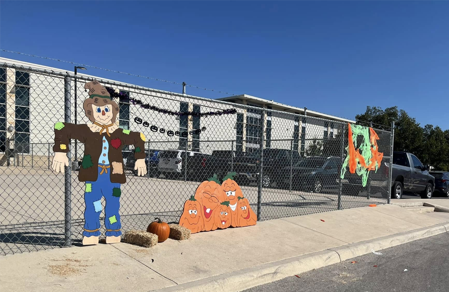 exterior of clemens high school beneath a cloudless sky of blue. chainlink fence is festooned with a cartoonish scarecrow and pumpkins.