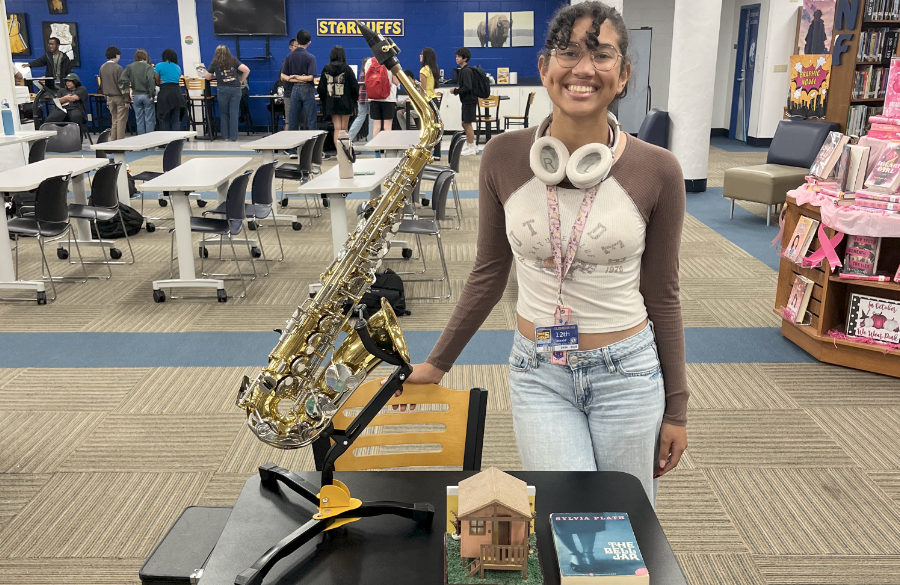 a high school girl with glasses & headphones smiles at the camera while standing behind a table featuring an alto saxophone, a model of a house, and sylvia plath's "the bell jar." in background is a high school library with several people gathered around similar exhibitions.