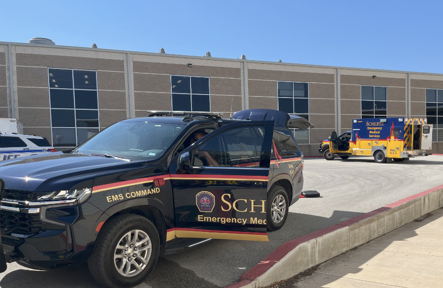 emergency services suv sits in foreground with driver's door open. an ambulance & police suv are in background before high school exterior beneath a blue sky
