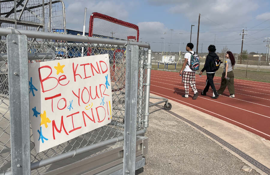 three high school students walk on an outdoor track beneath a morning sky. a sign in foreground reads "be kind to your mind!"