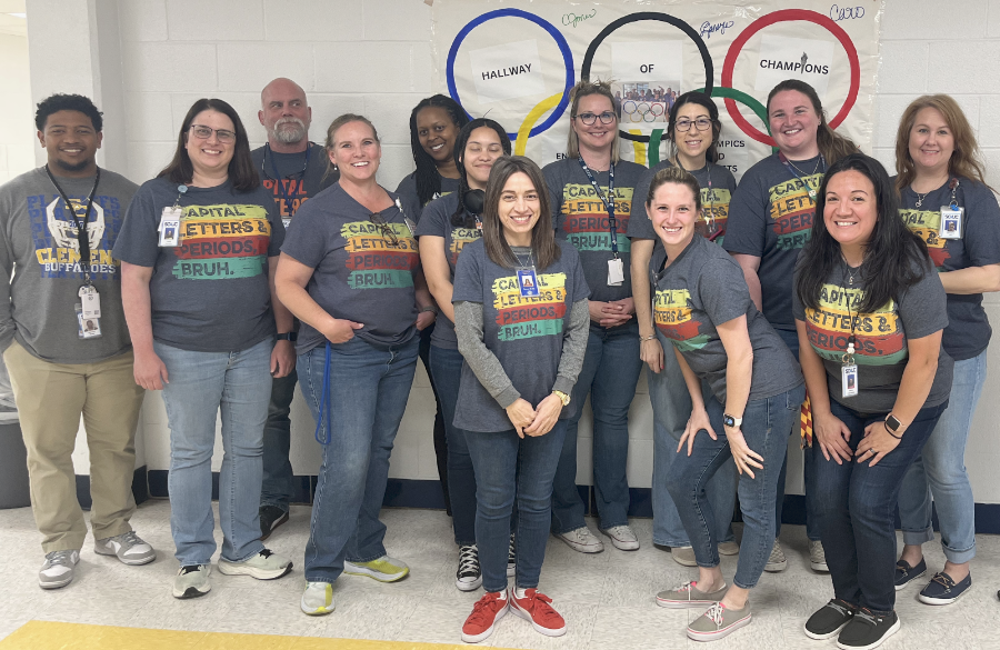 a cadre of teachers stand smiling before an olympic ring banner in a high school hallway, wearing matching shirts which read "capital letters & periods, bruh."