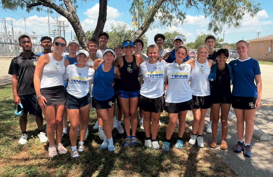 high school tennis players stand beneath the shade of a tree on a grassy field in athletic attire, arms linked & smiling