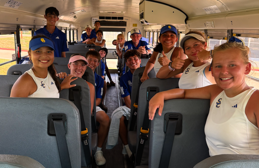 tennis players in white & blue uniforms sit on a school bus, smiling & giving thumbs up to the viewer
