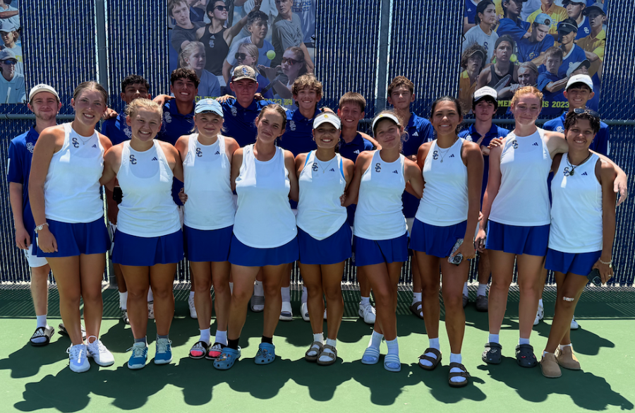 high school tennis players in blue & white uniforms stand before tennis graphics on a tennis court beneath a sunny sky
