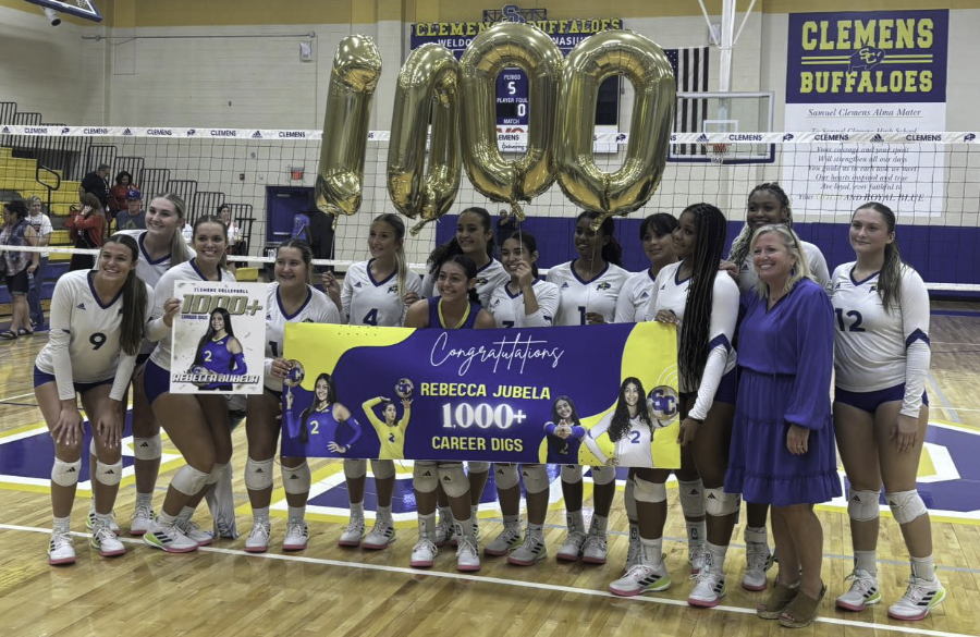 high school volleyball players stand midcourt in a gym before a volleyball net holding numbered balloons for "1000" and a banner reading "congratulations rebecca jubela 1000+ career digs"