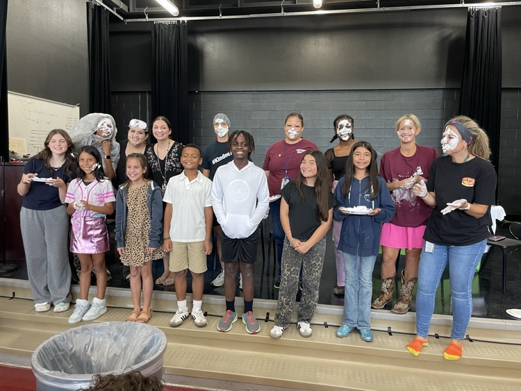 Students and teachers lined up on the stage posing with pie on their face