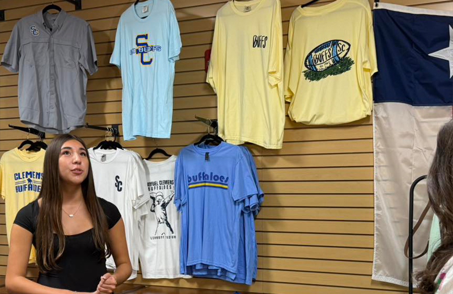 a high school student stands before a slatted wall with high school t-shirts on display behind her. she speaks to an out-of-frame news reporter