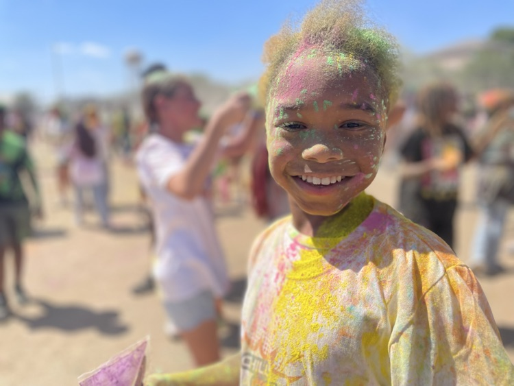 student smiling at the camera, covered in colored powder outside on the field with their peers in the background 