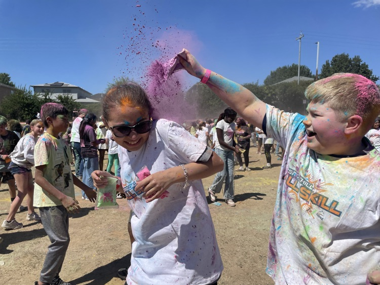 student smiling at the camera, covered in colored powder outside on the field with their peers in the background 