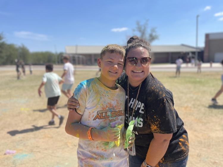 student smiling at the camera, covered in colored powder outside on the field with their peers in the background 