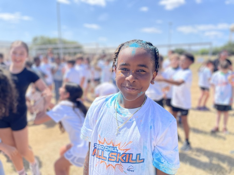 student smiling at the camera, covered in colored powder outside on the field with their peers in the background 