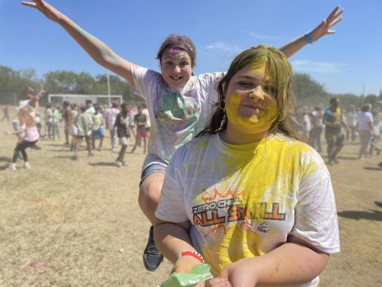 student smiling at the camera, covered in colored powder outside on the field with their peers in the background 