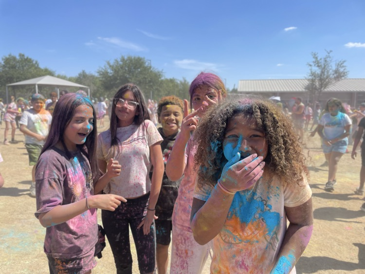 student smiling at the camera, covered in colored powder outside on the field with their peers in the background 
