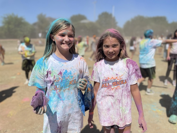 student smiling at the camera, covered in colored powder outside on the field with their peers in the background 