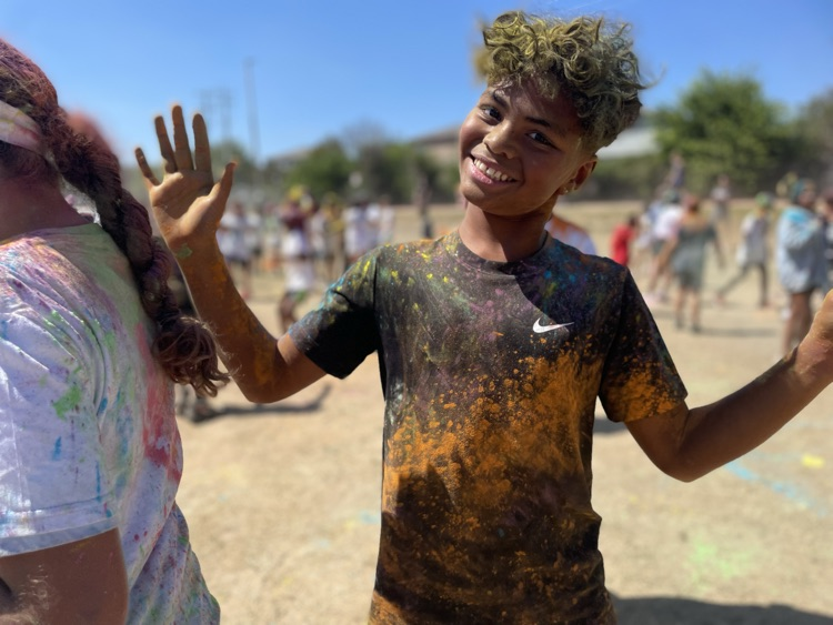 student smiling at the camera, covered in colored powder outside on the field with their peers in the background 