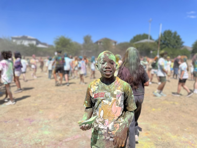student smiling at the camera, covered in colored powder outside on the field with their peers in the background 