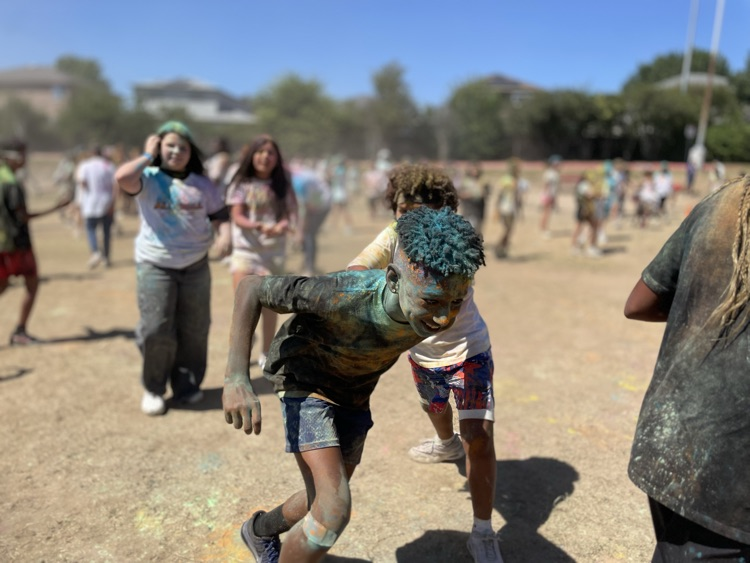 student smiling at the camera, covered in colored powder outside on the field with their peers in the background 