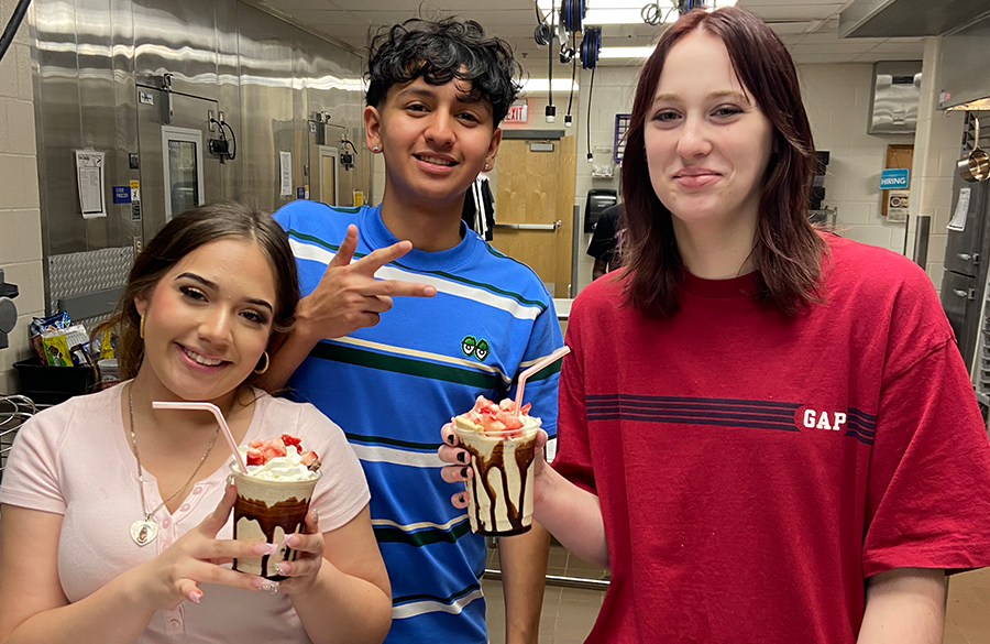 a trio of high school students stand in a culinary arts classroom holding gourmet milkshakes topped with fresh fruit & straws
