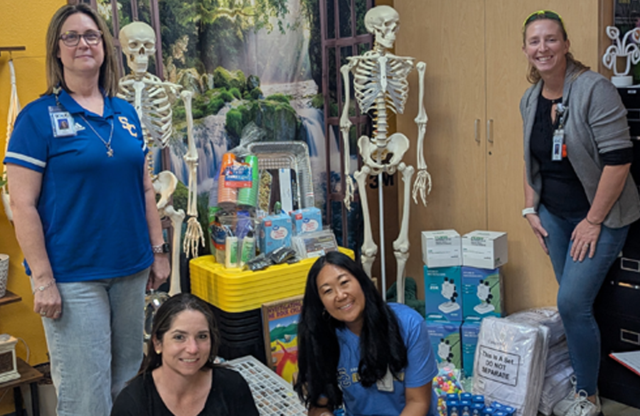 four science teachers crowd around several supplies for use in a science classroom, including two skeleton replicas, bins, and so on