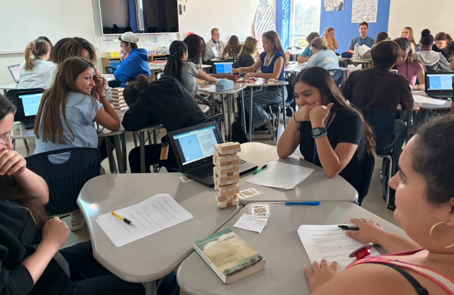 a high school classroom features desks clustered in groups of three, each with a laptop and jenga block game. in foreground, three female students stare in awe at a precarious tower as they respond to questions on review papers.